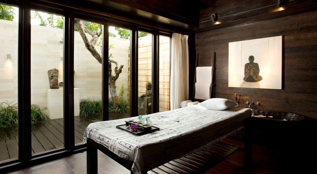 Modern spa room with massage table, dark wood walls, and large sliding doors overlooking a garden