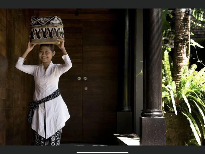 Woman in white tunic balancing a carved basket on her head beside a wooden doorway.