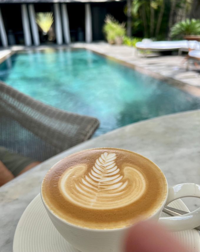 Latte with leaf latte art on a table beside a turquoise pool in a resort courtyard