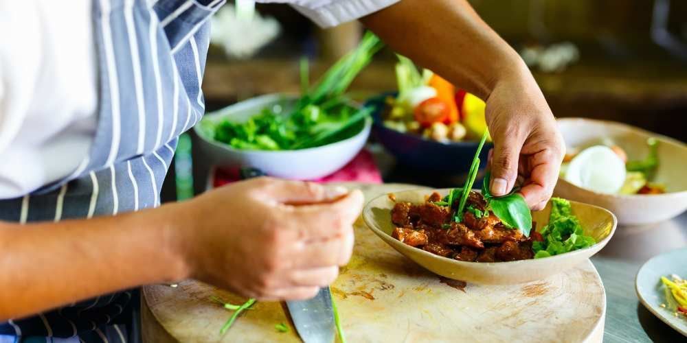 Hands plating a green salad and roasted meat at a dining table