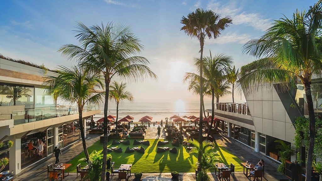 Sunlit tropical courtyard with palm trees, people dining, and ocean view at sunset
