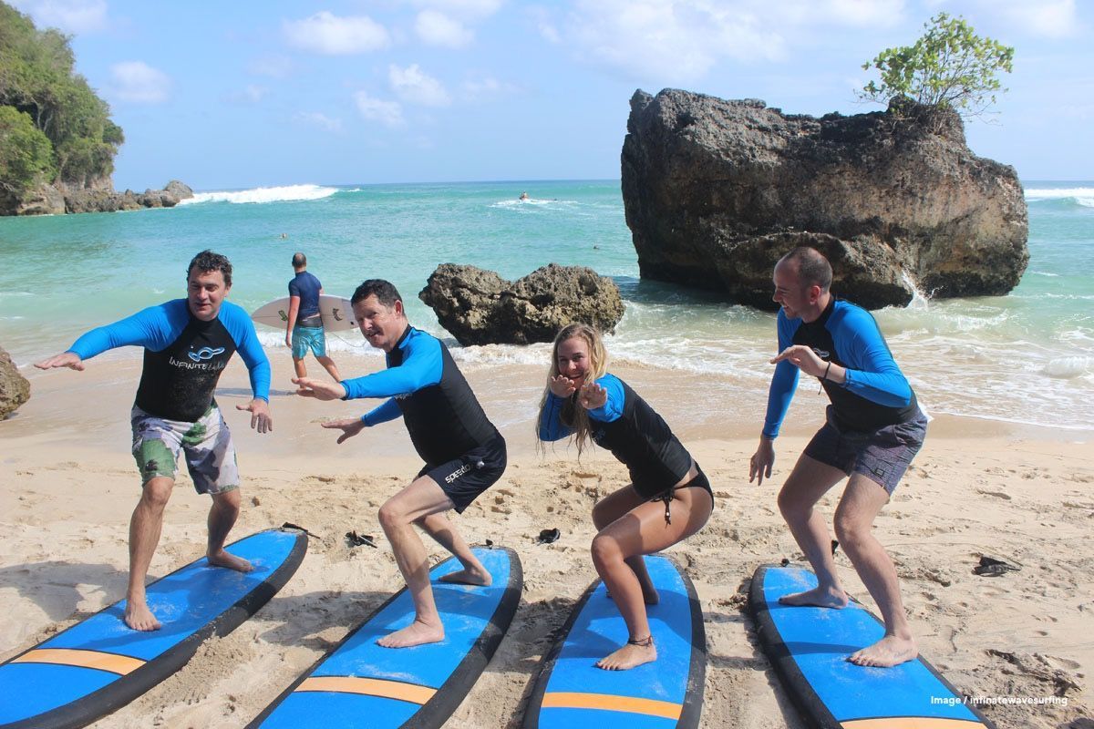 Surf lessons Four people in blue shirts balance on blue surfboards on a sandy beach by the ocean.