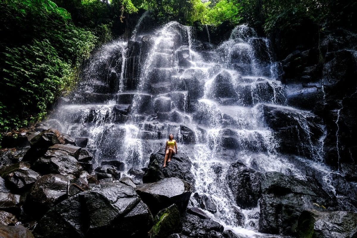 Person standing on rocks beneath a cascading waterfall in a lush green forest