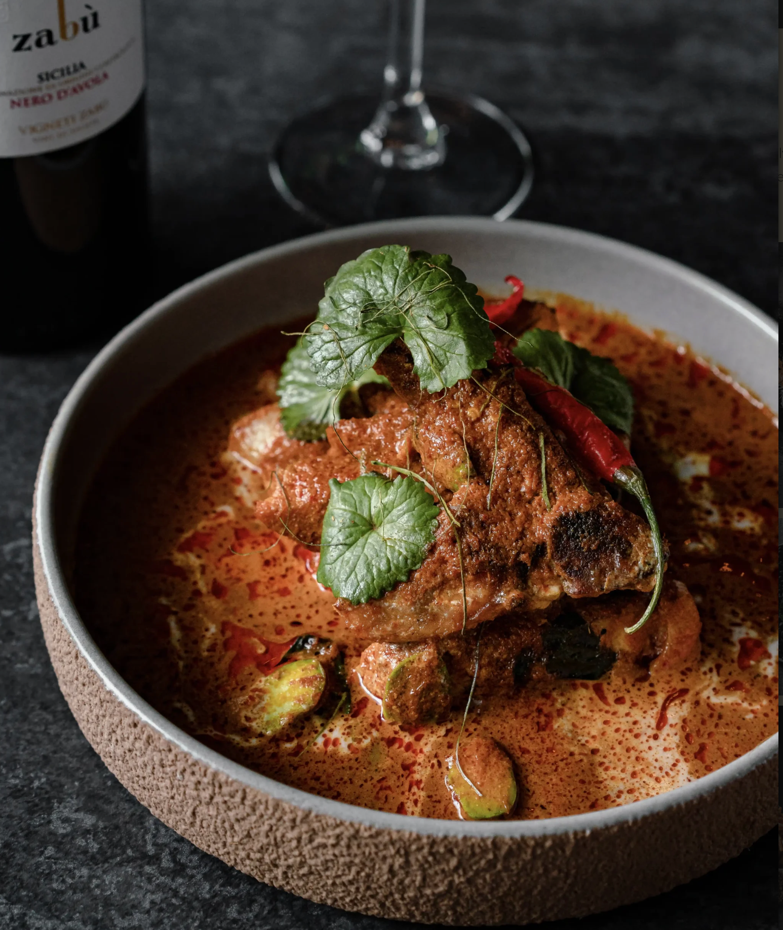 Bowl of creamy curry with herb garnish on a dark table, beside a wine glass and bottle