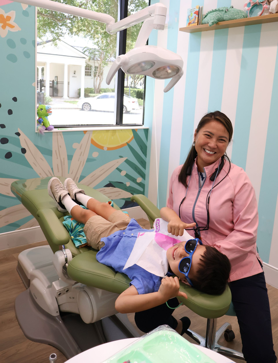 A child is sitting in a dental chair while giving thumbs up and smiling at the camera along with the dentist