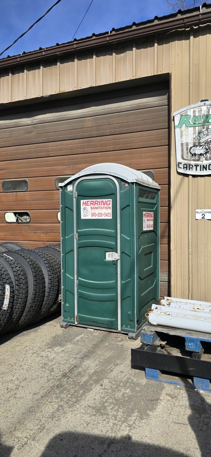 A green portable toilet is sitting in front of a garage next to a pile of tires.