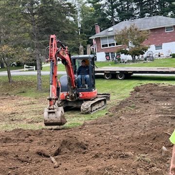 A man is driving an excavator on a dirt field in front of a house.