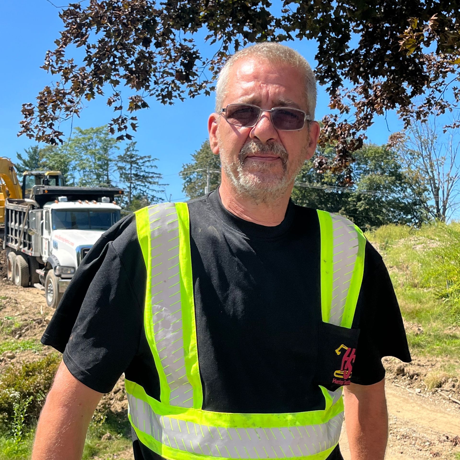A man wearing a black shirt and a yellow vest is standing in front of a construction site.