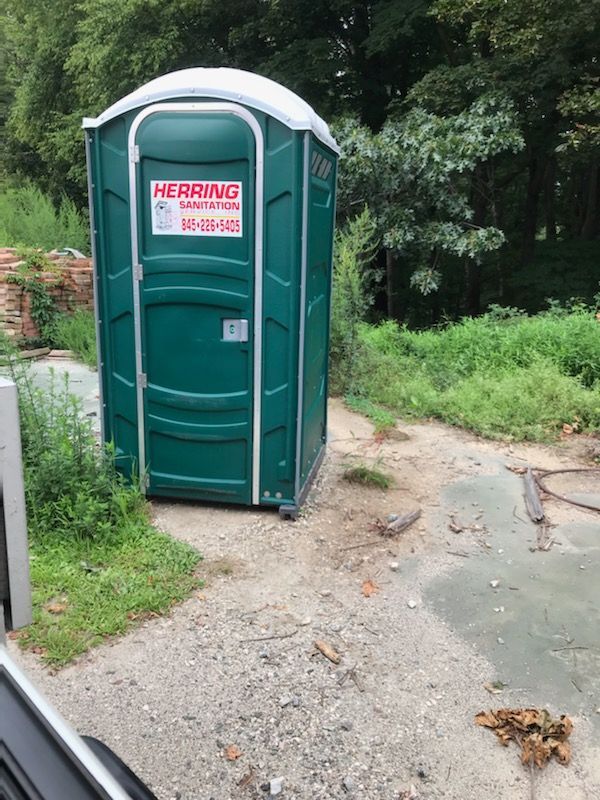 A green portable toilet is sitting on the side of a dirt road.