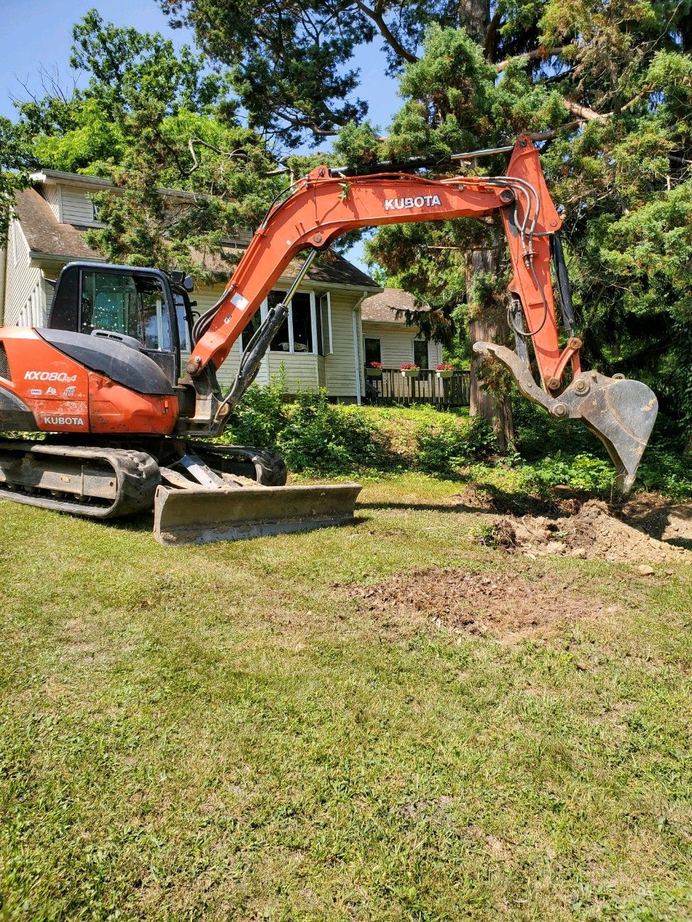 An excavator is digging a hole in the grass in front of a house.