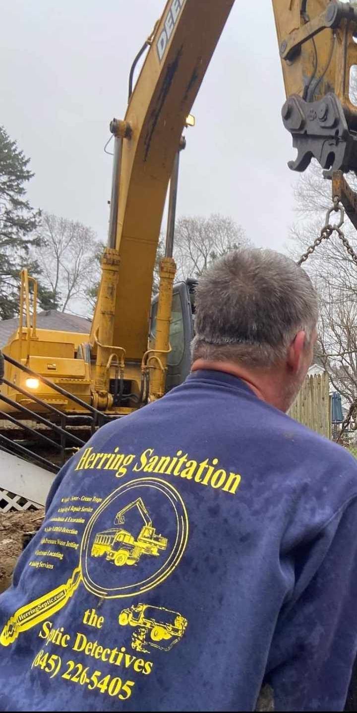A man in a blue shirt is standing in front of a yellow excavator.