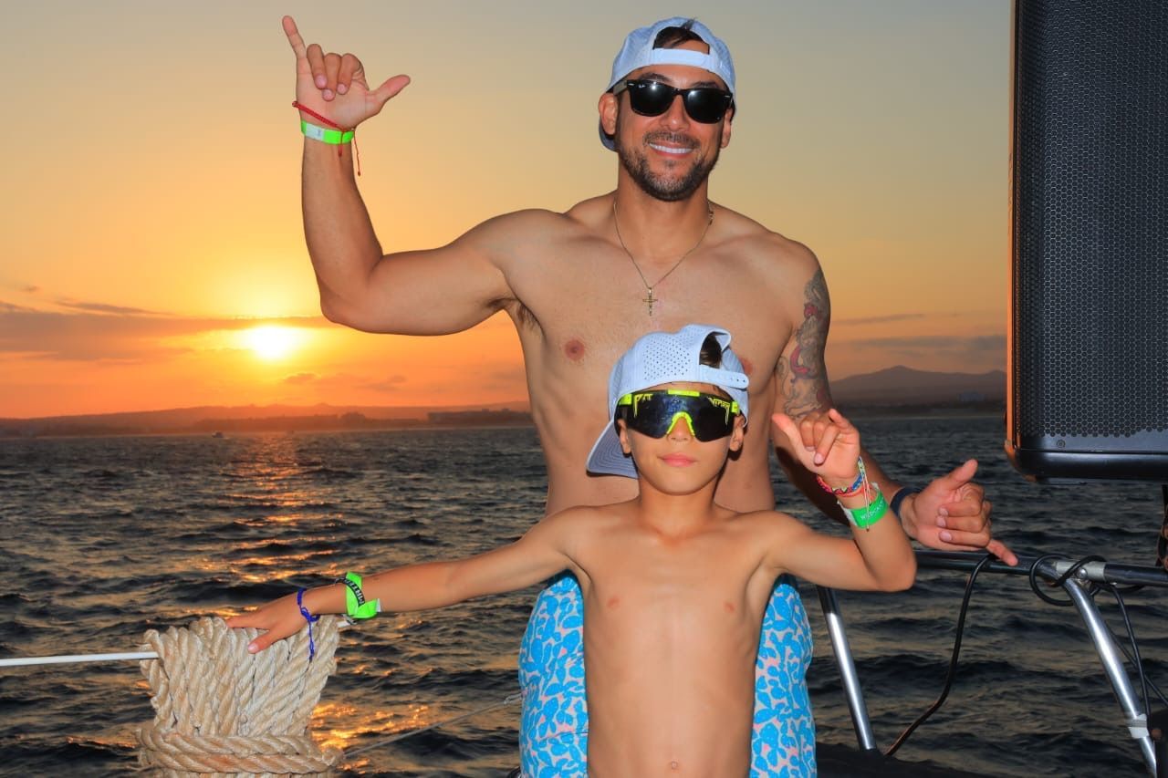 Two people posing on a boat at sunset, wearing sunglasses and festival wristbands.