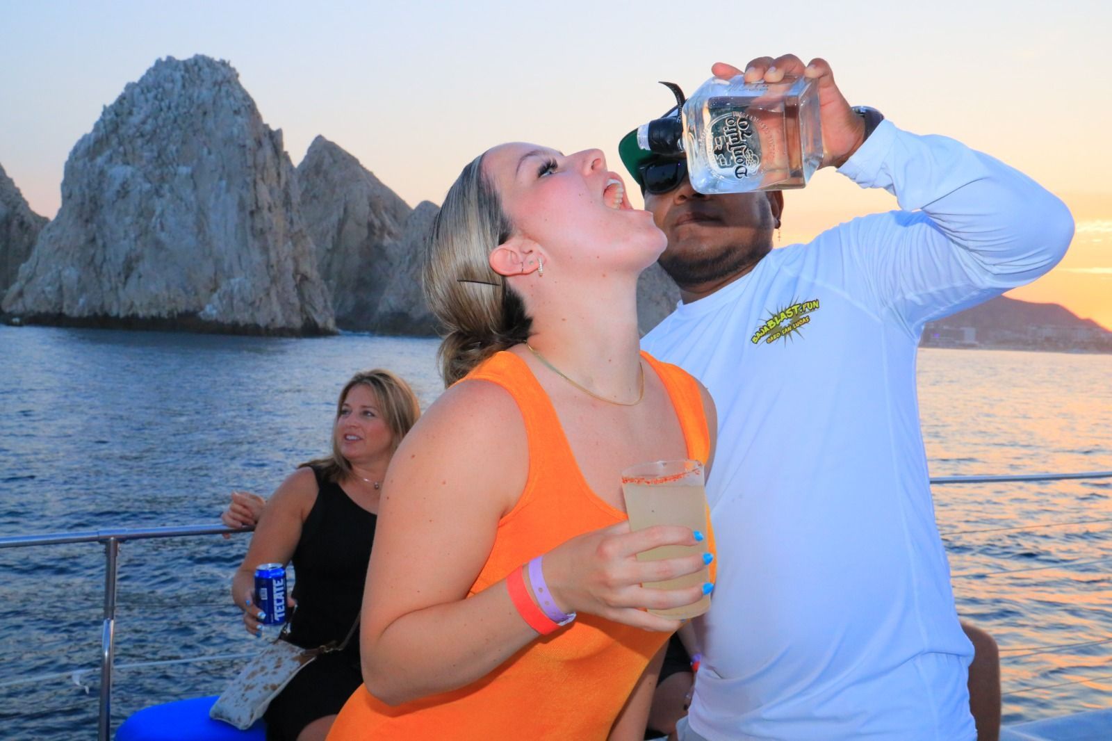 People on a boat by a rocky coast, one drinking from a bottle at sunset.