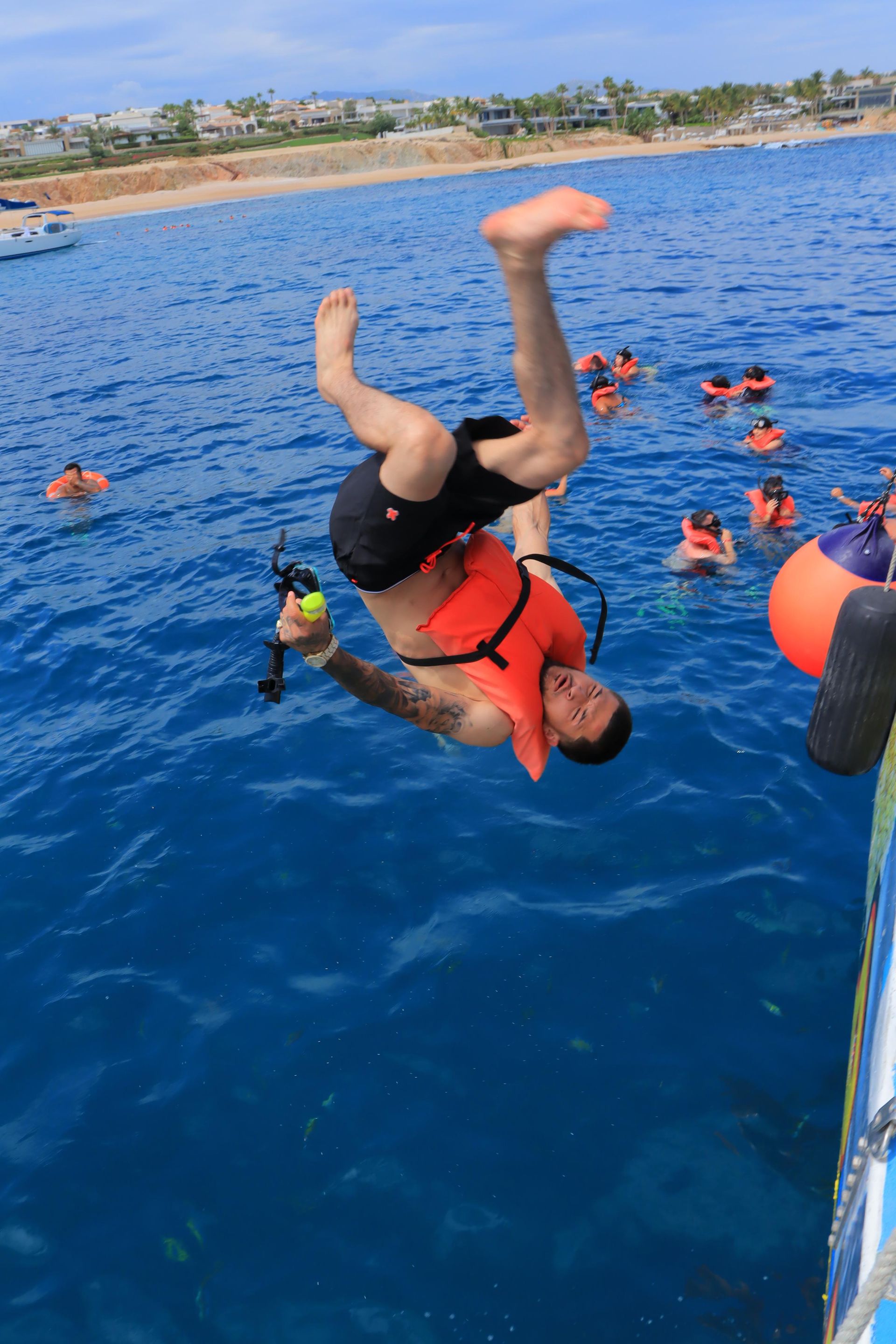 Person in orange life vest flipping backward into blue water near snorkelers by a boat.