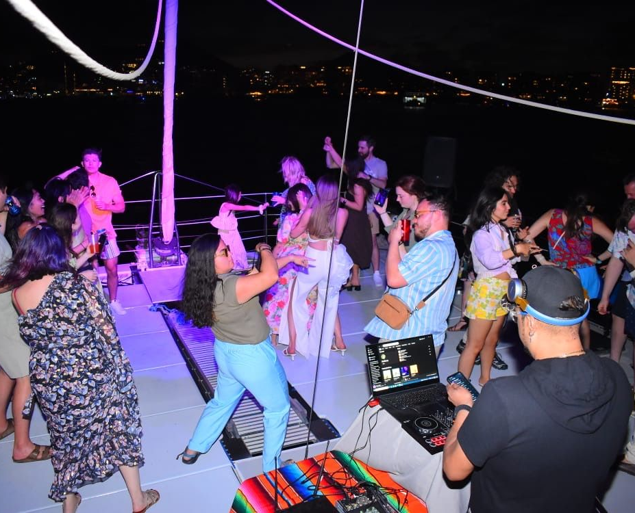 Sunset Cruise Tour Cabo San Lucas People dancing on a lit outdoor deck at night, with colorful lights and a distant shoreline.