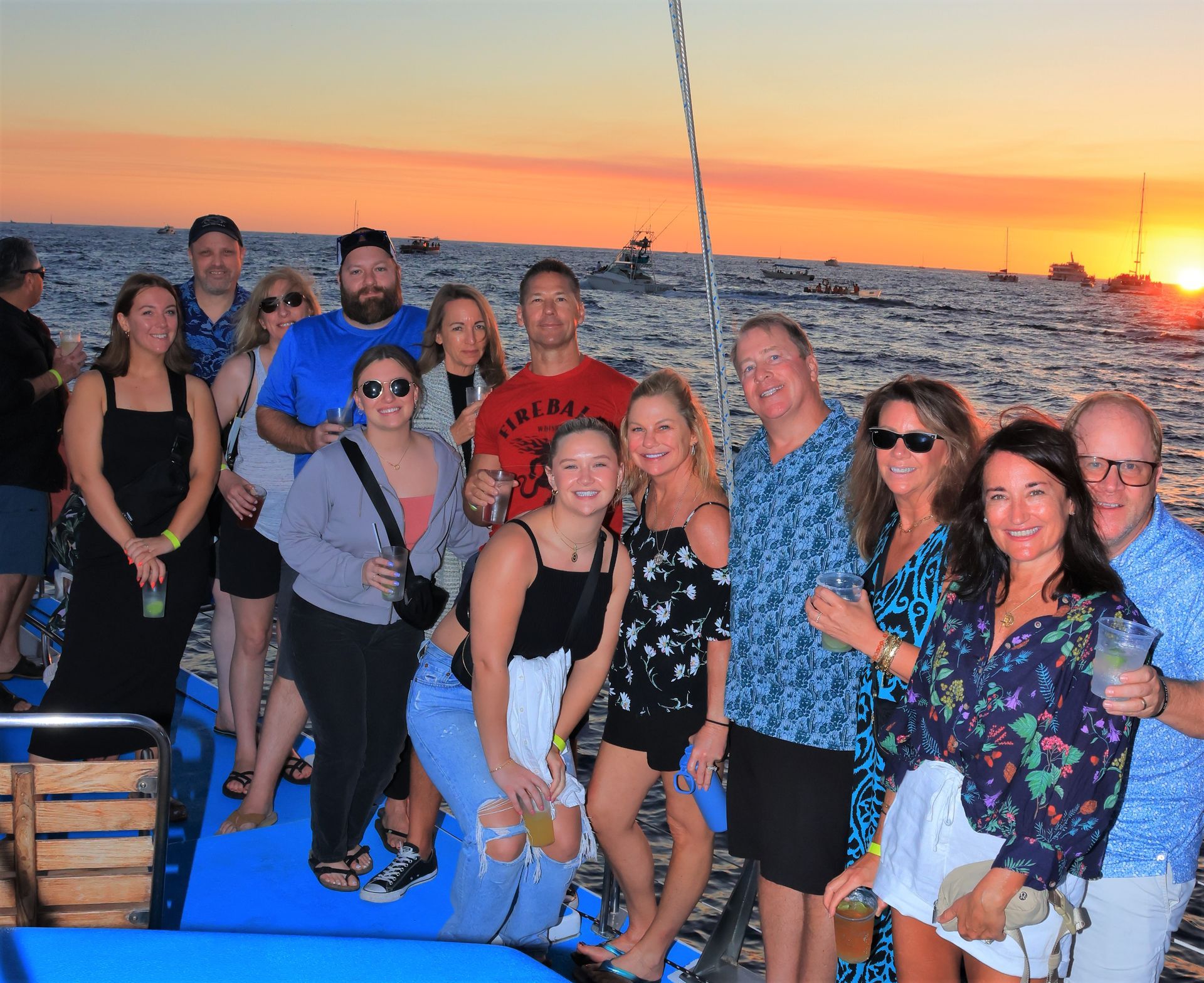 Group of people smiling on a boat at sunset on the water, with an orange sky and distant city lights.