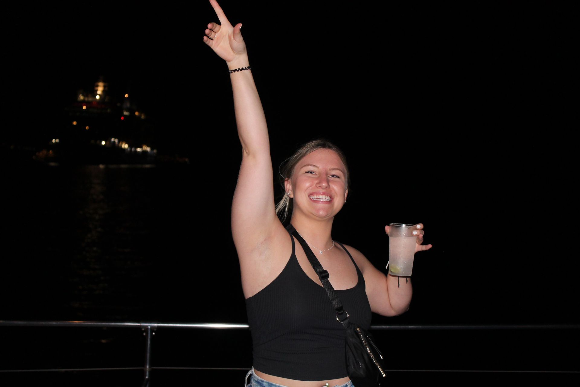 Smiling person in a black top raises one arm and holds a drink at night by the water.