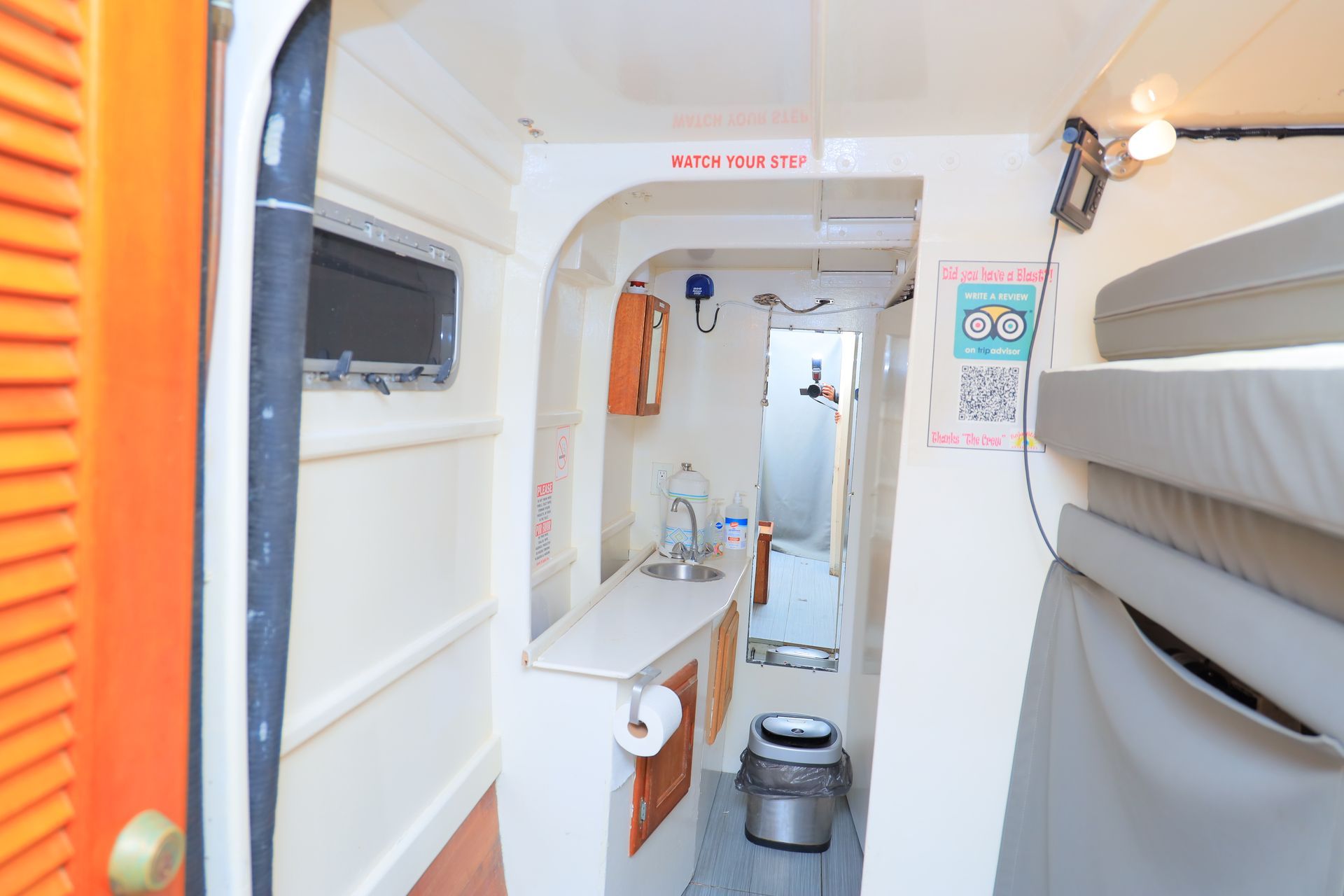 Narrow white boat corridor with orange door, shelves, sink, and equipment onboard