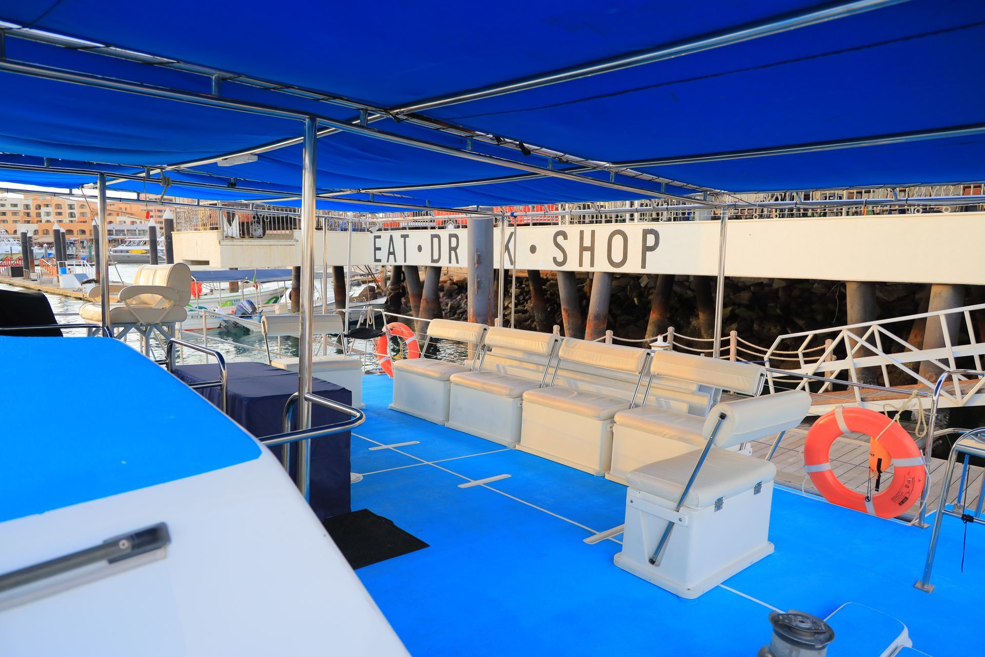 Open-air boat deck with blue flooring, shop counters, railings, and a life ring under a canopy