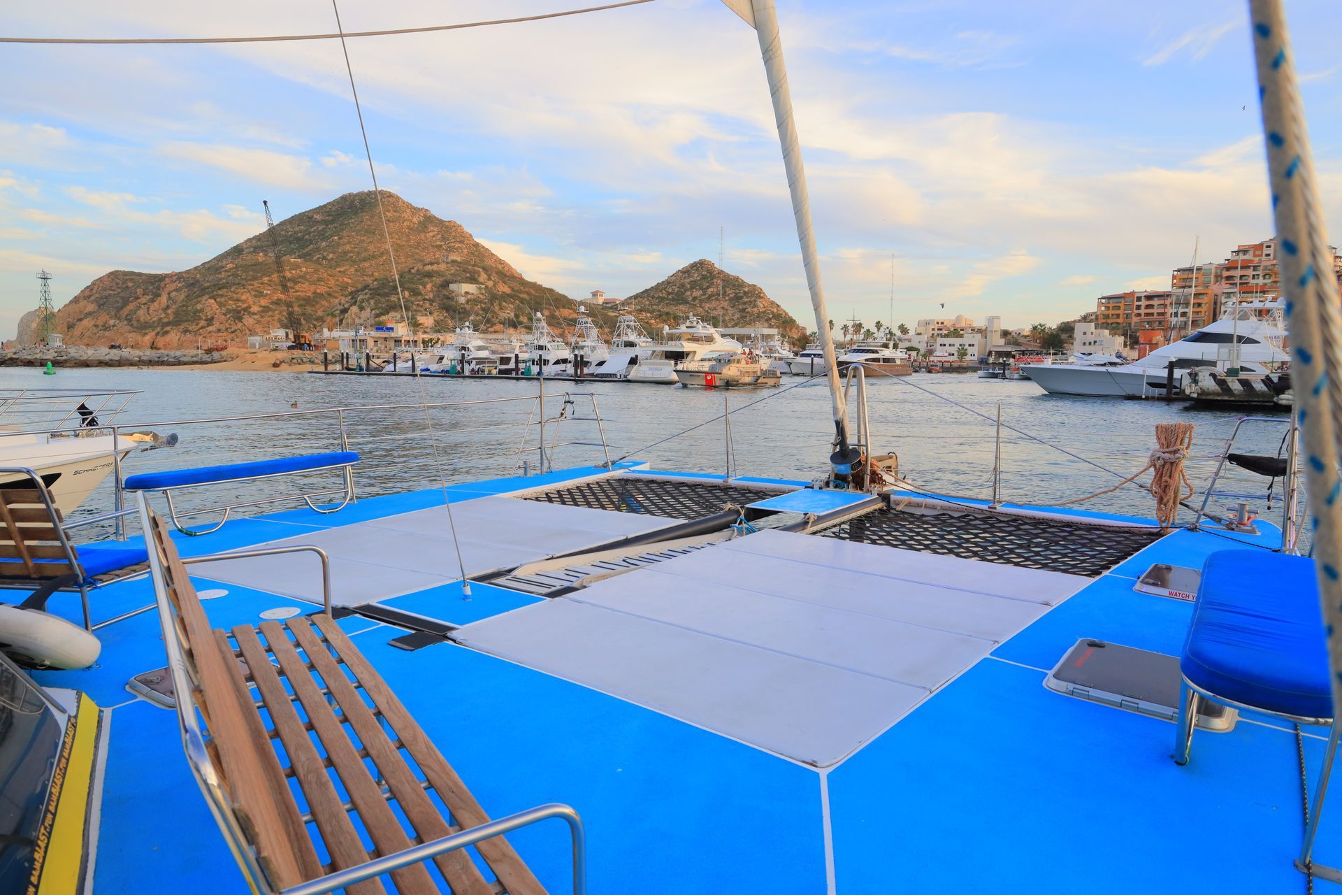 Blue-deck sailboat with wooden bench seating in a harbor, hills and boats in the background