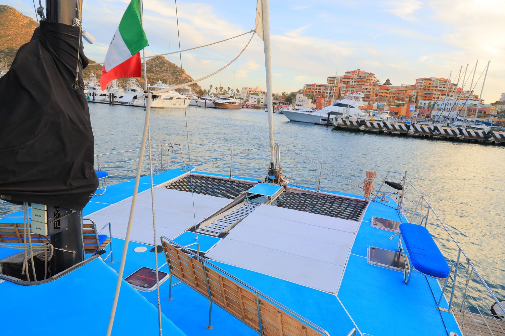 Blue sailboat deck overlooking a harbor with Italian flag and a waterfront town at sunset