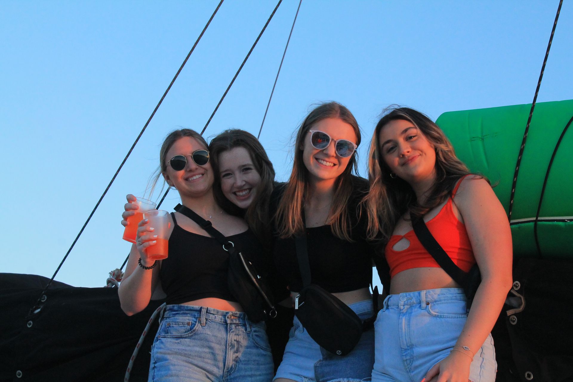 Four friends smiling outdoors, wearing sunglasses and casual clothes at sunset with blue sky and colorful background