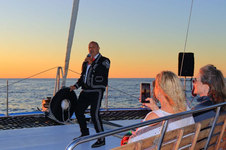 Singer performing on a sailboat at sunset, with two people recording from the deck.