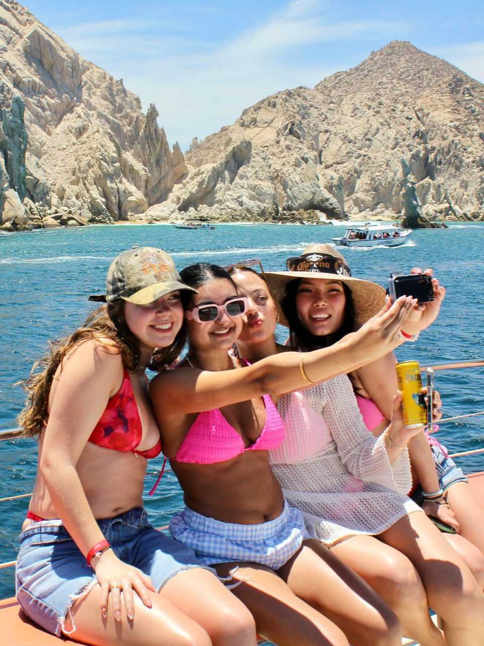 Four friends smiling on a boat, taking a selfie with rocky sea cliffs in the background.