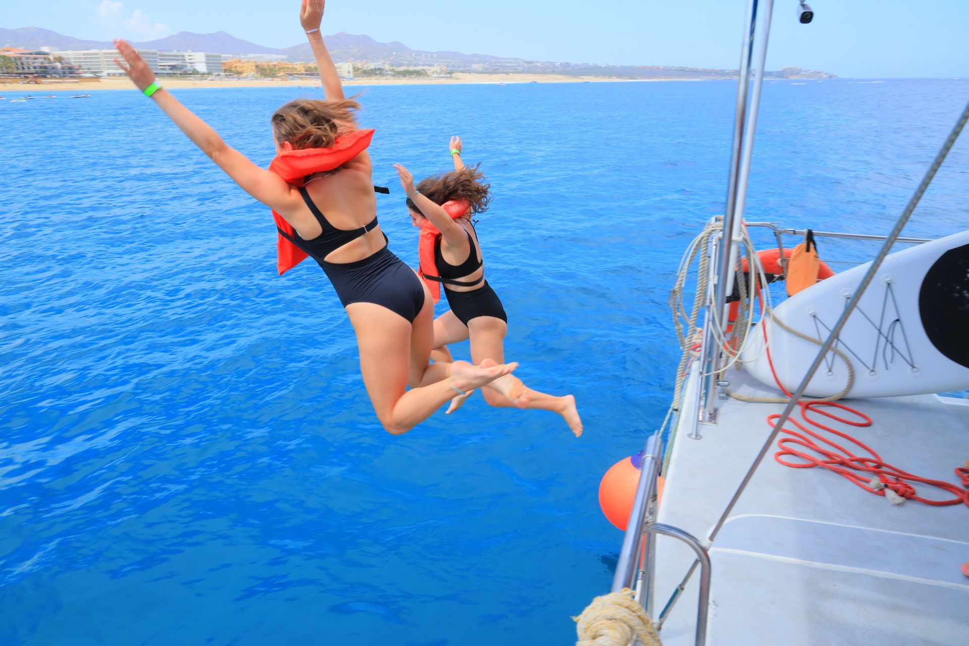 Two people leap from a sailboat into bright blue water near a coastline.