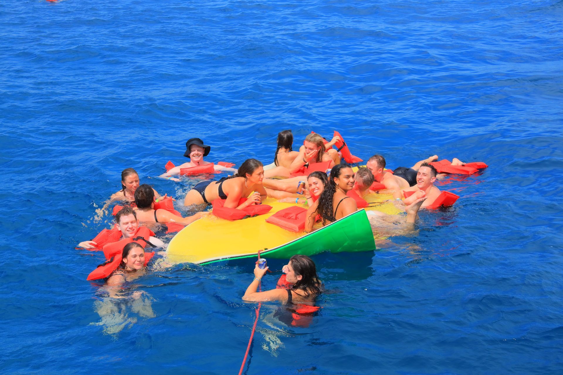 Group floating on a colorful inflatable raft in blue water, with a person holding a rope in front.