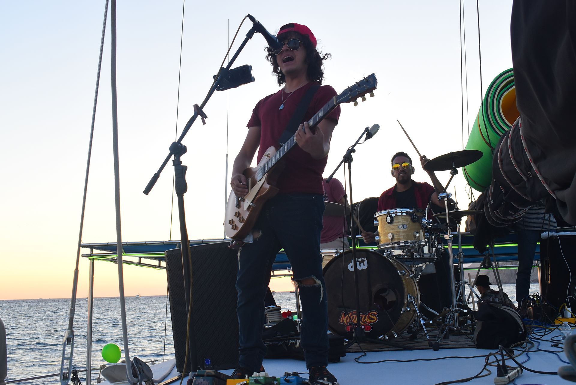 Musician playing guitar on an outdoor stage by the water at sunset, with drummer behind.