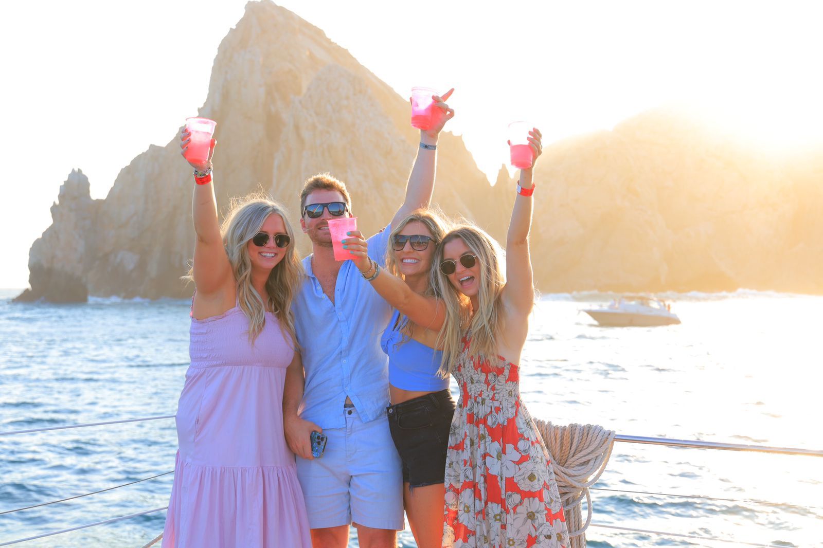 Four friends holding pink drinks and smiling on a sunny beach at sunset, with rocks and ocean behind them