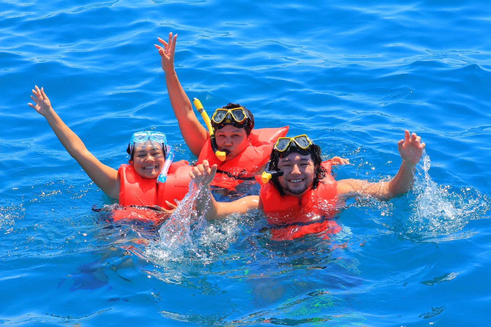 Three snorkelers in red life vests wave in bright blue ocean water