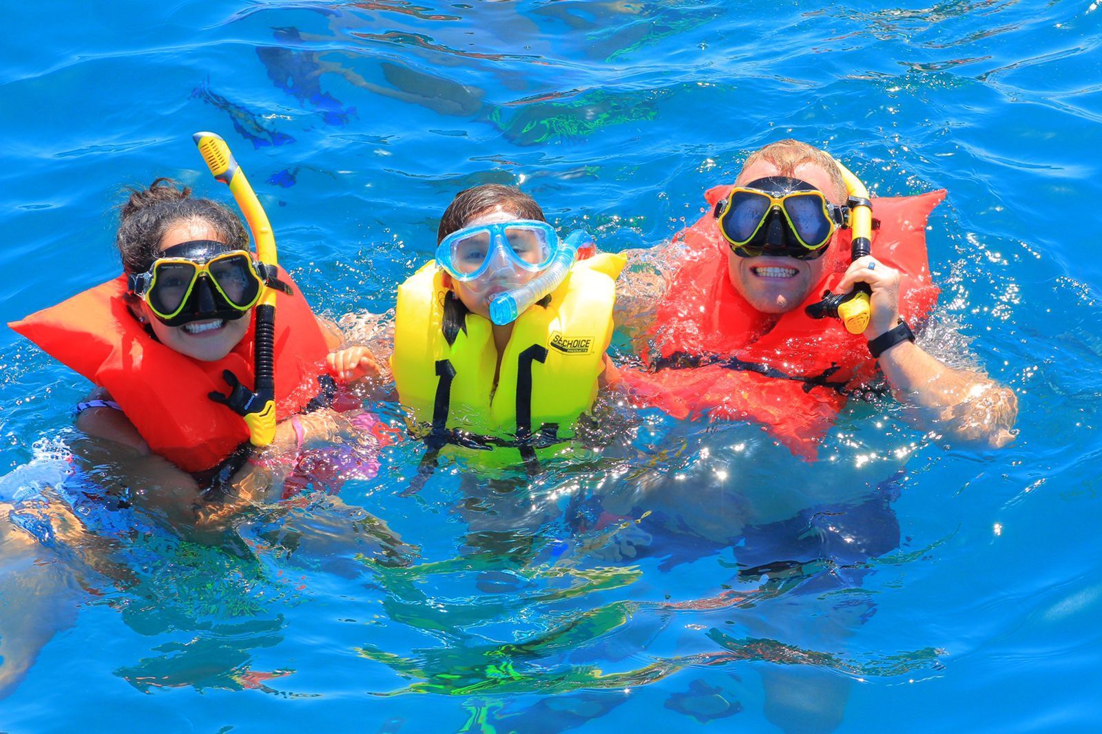 Three snorkelers in colorful life vests floating in bright blue ocean water