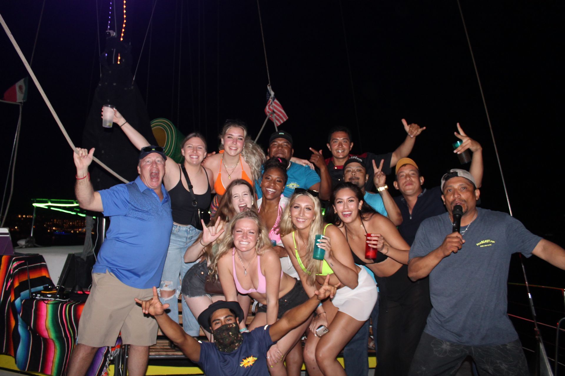 Group of people posing and cheering on a boat at night, holding drinks and wearing leis.