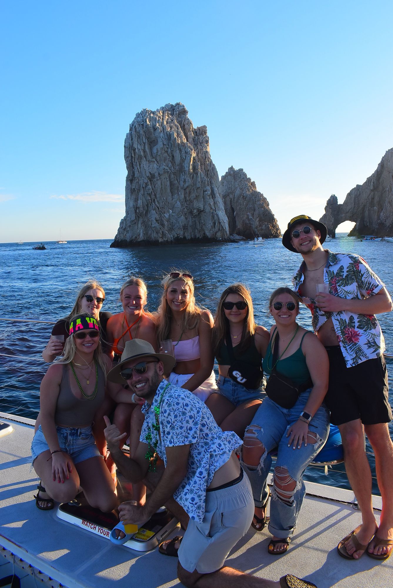 Group posing on a boat deck by the sea, with a large rock formation and blue sky in the background.