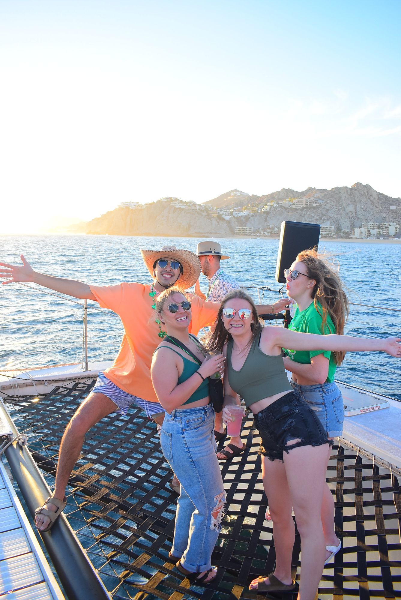 Four friends pose on a boat deck at sunset, arms outstretched with blue water and mountains behind them.