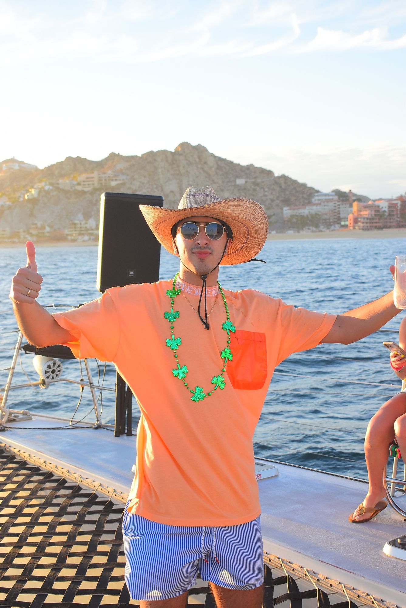 Person in orange shirt and straw hat posing on a boat with arms outstretched near the coast.