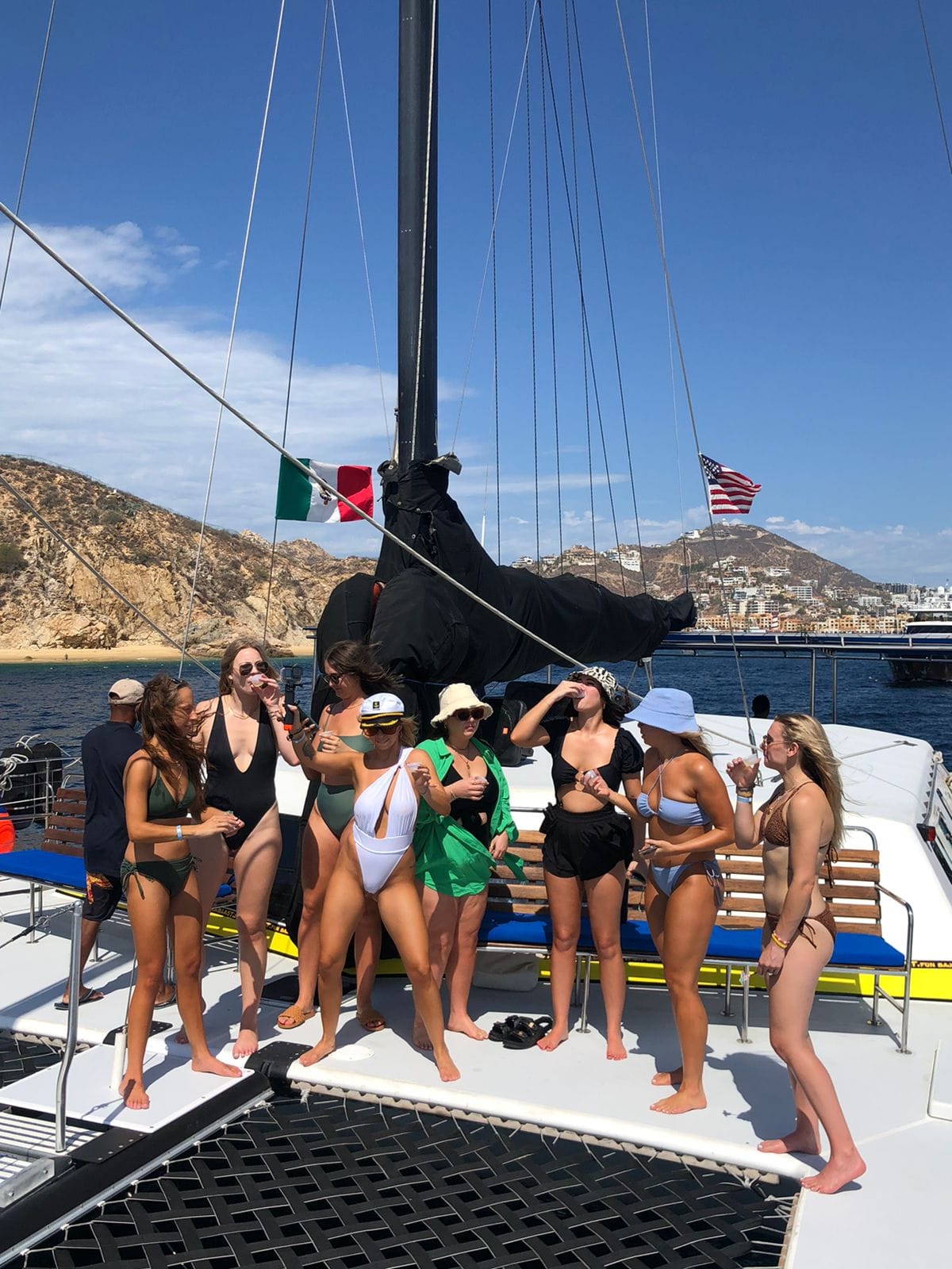 People posing on a catamaran deck near the sail, with Italian and American flags, on a sunny sea day