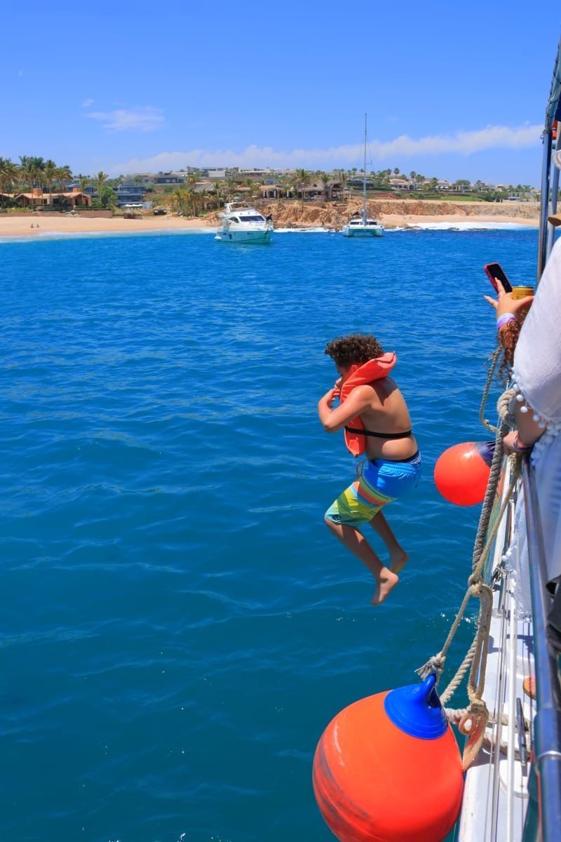 Person jumping off a boat into bright blue water near a sandy beach with a small boat in the distance