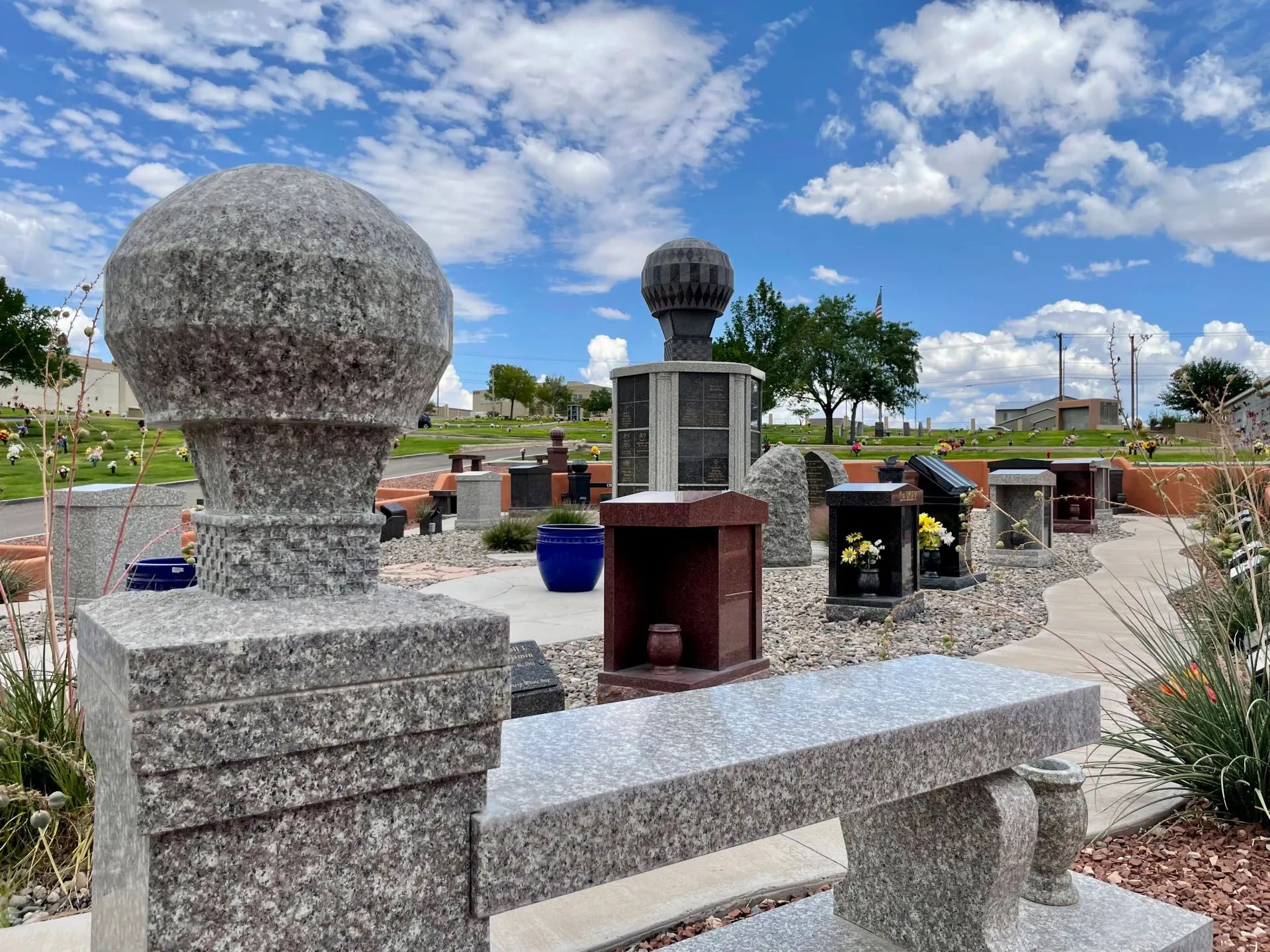 Granite bench and column in a cemetery, with headstones under a blue sky with clouds.