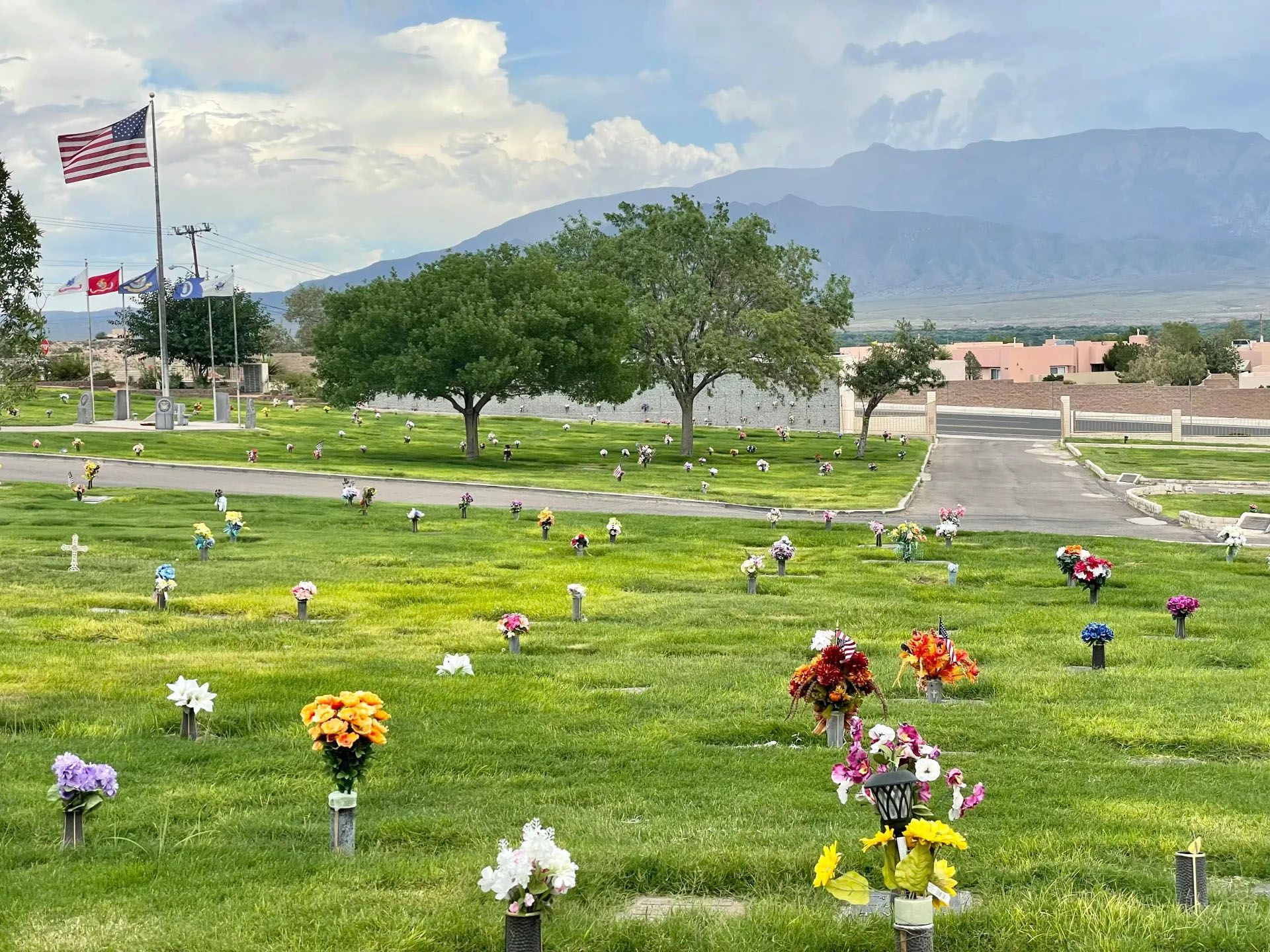 Cemetery with green grass, headstones, colorful flowers, and a mountain backdrop under a cloudy sky.