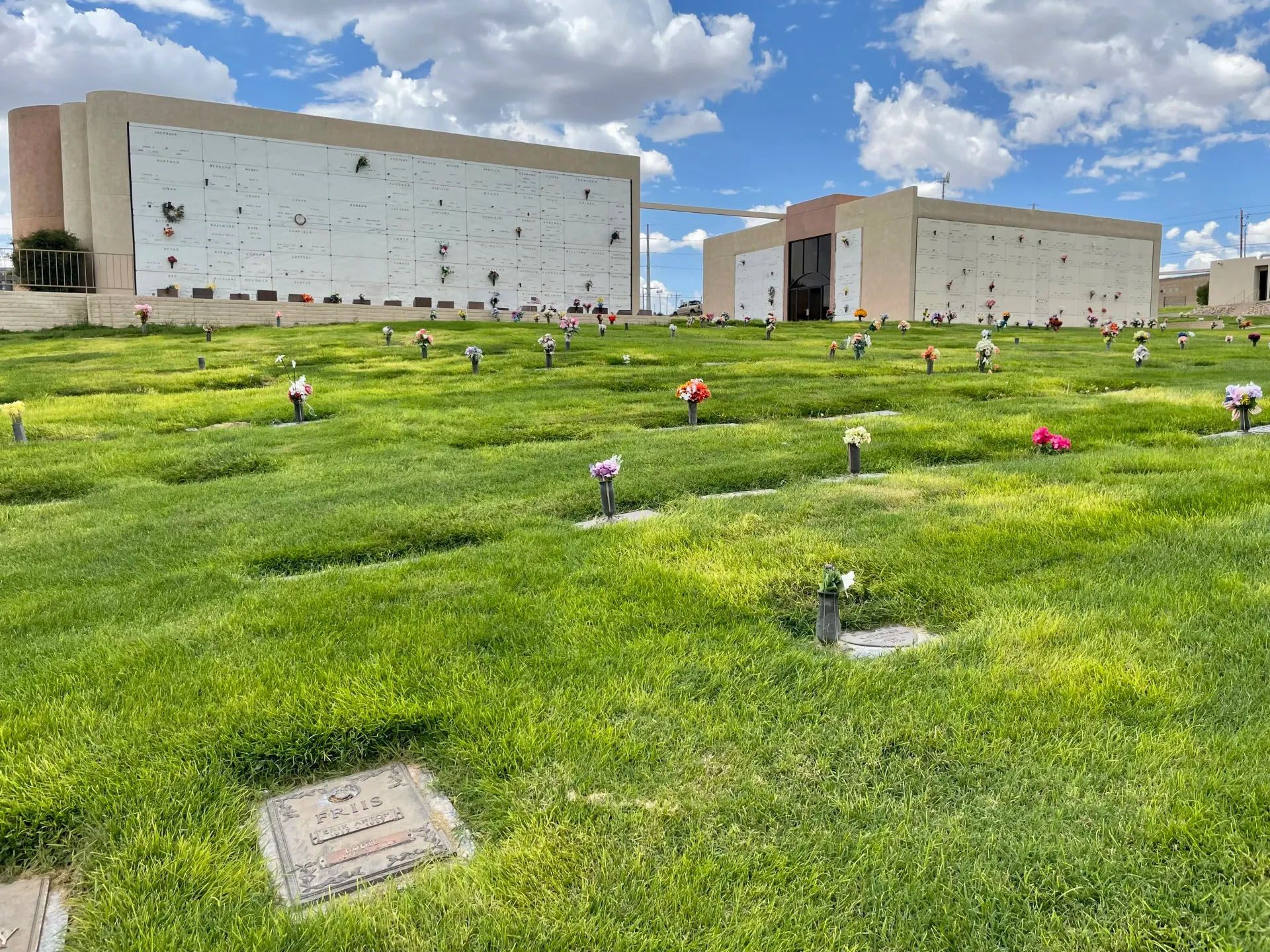 Cemetery with green grass, rows of markers, and beige memorial buildings under a cloudy sky.
