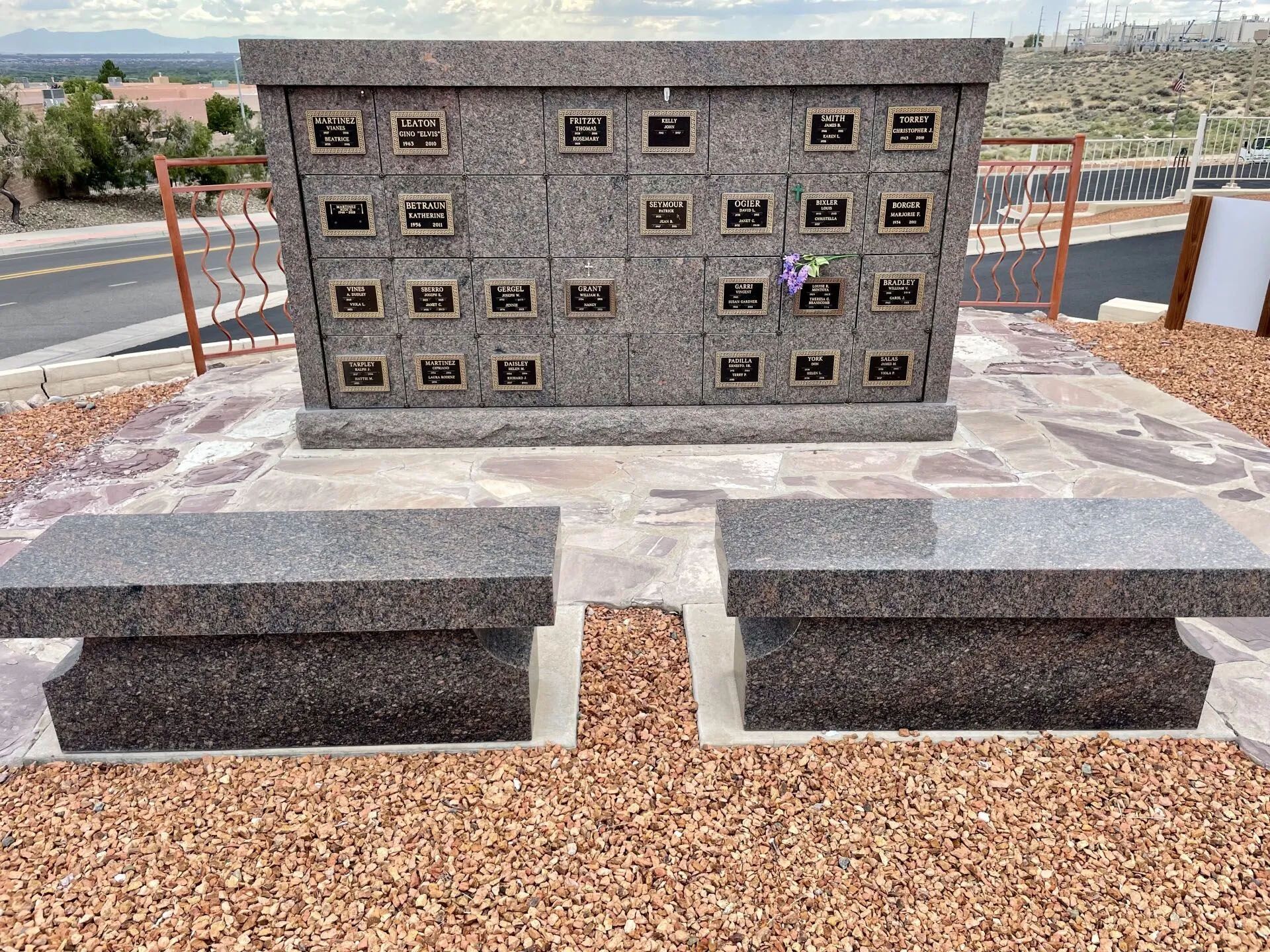 Granite columbarium with bronze plaques and two benches in a cemetery setting with gravel.