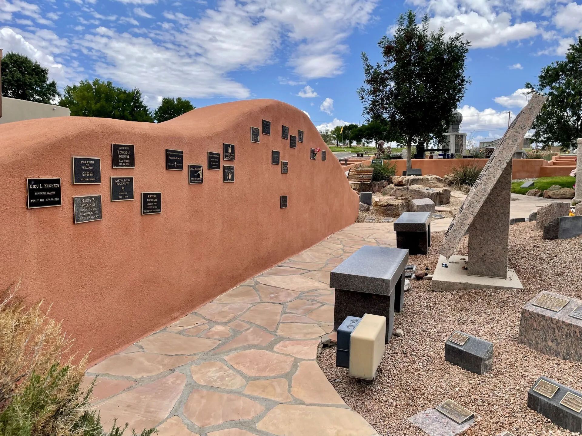 A memorial garden with a terracotta-colored wall displaying plaques, stone benches, and sculptures under a blue sky.