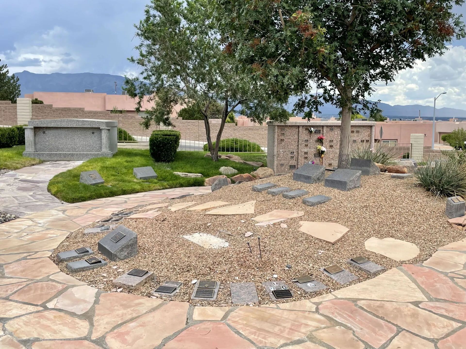 Cemetery with headstones, trees, and columbarium. Gravel and stone path. Cloudy sky and distant mountains.