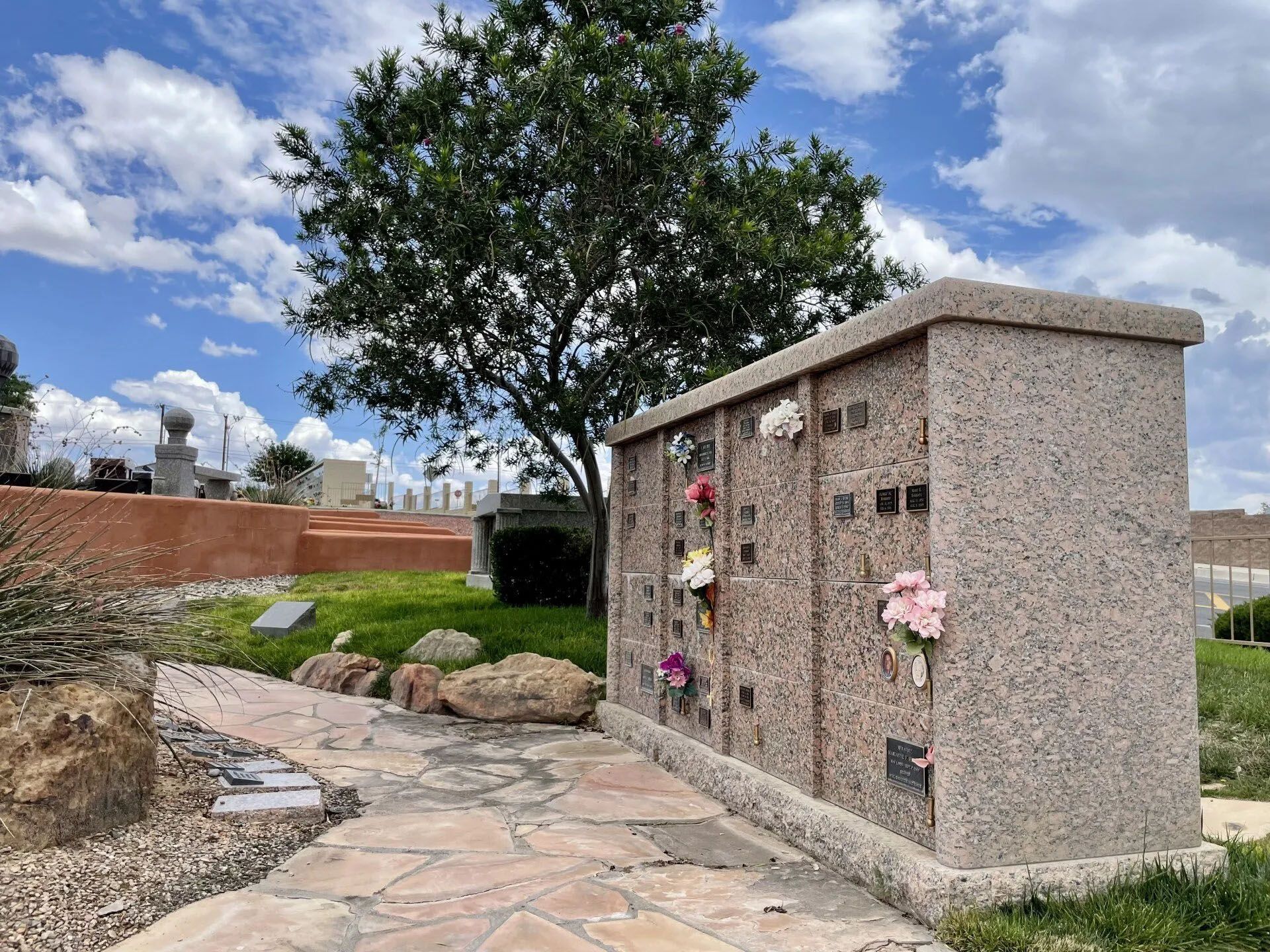 A stone columbarium with floral arrangements in a sunny cemetery setting.