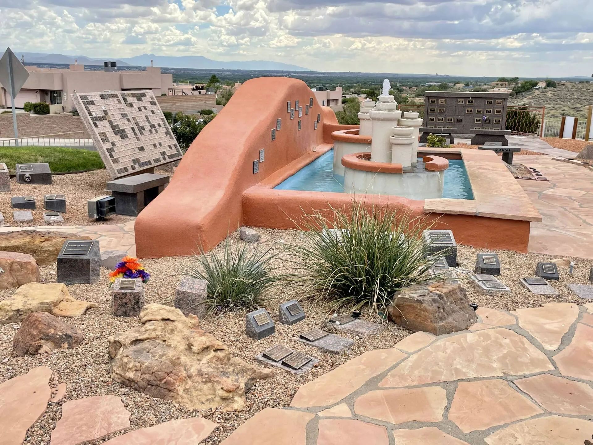 Terracotta-colored water fountain with tiered basins, surrounded by small headstones and desert landscaping.