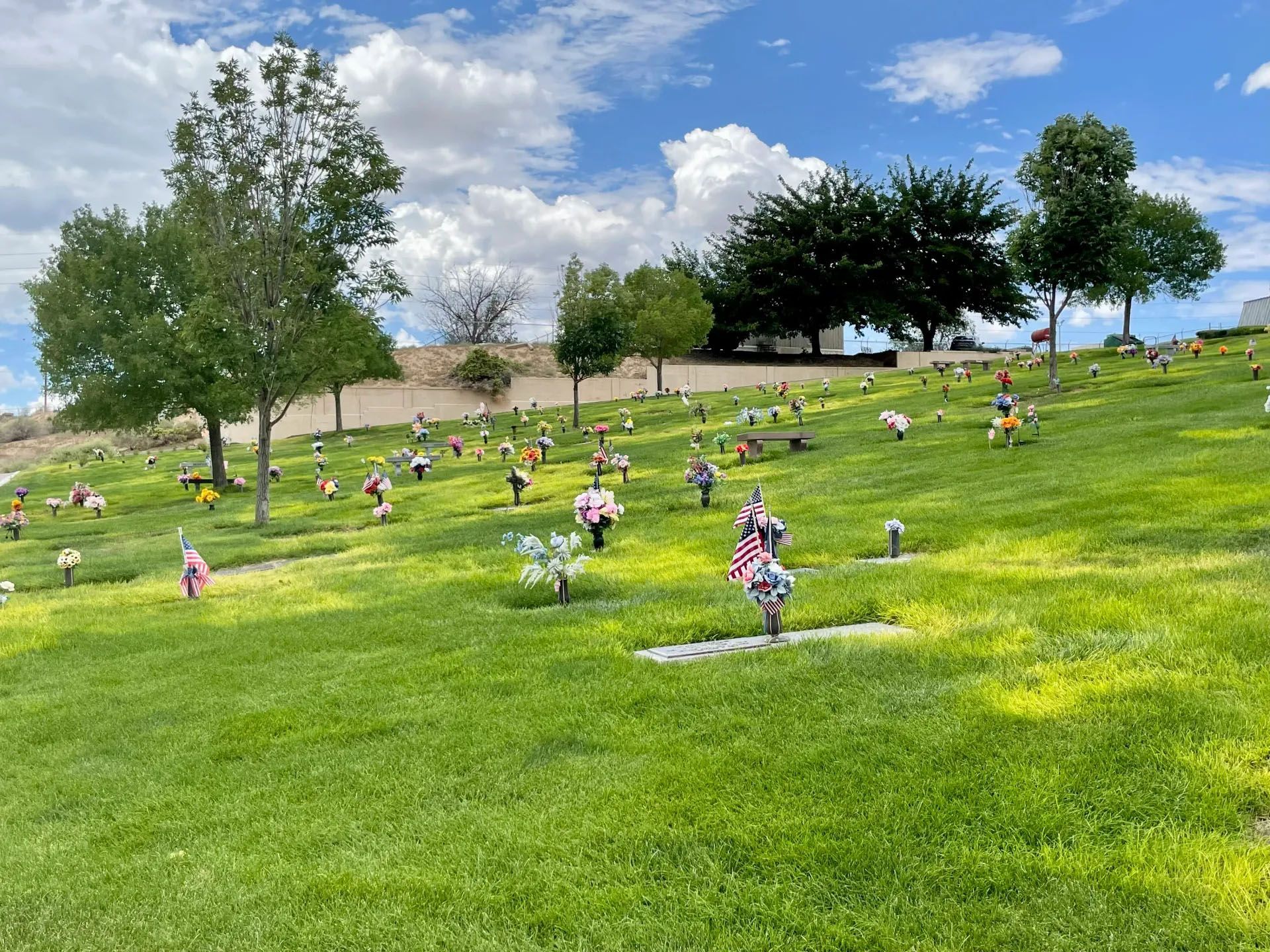 Rolling green hillside cemetery with headstones, flowers, and trees under a partly cloudy sky.