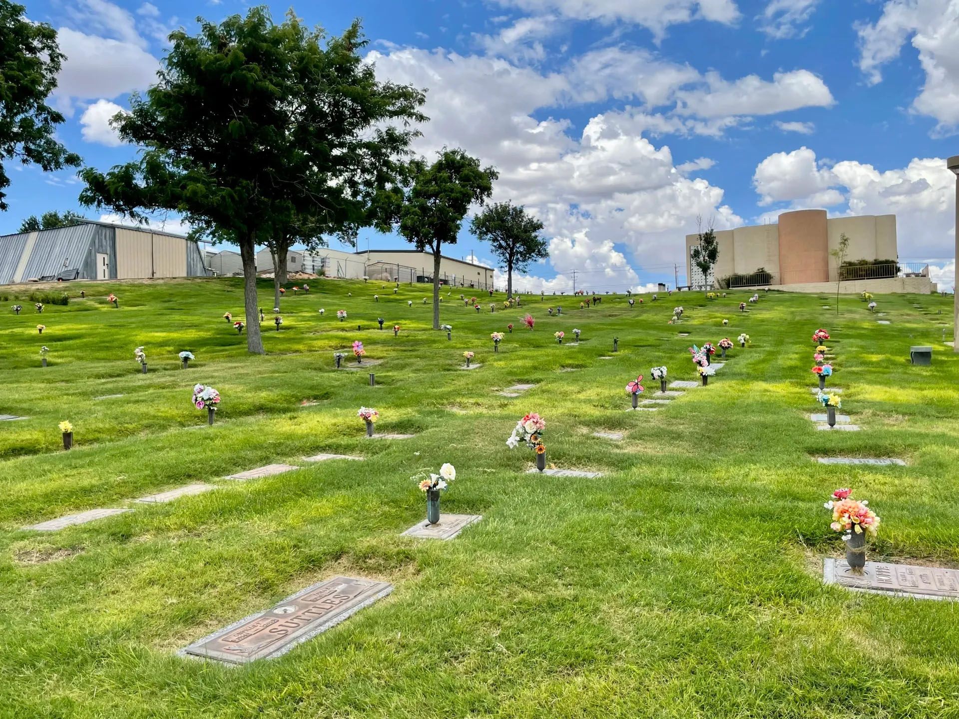 Cemetery on a grassy hill with headstones, flowers, trees, and buildings against a cloudy sky.